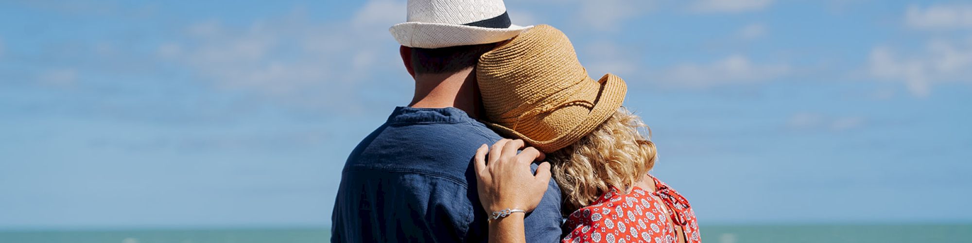 A couple in hats is embracing while looking at the ocean from a railing, under a sunny sky with scattered clouds, creating a peaceful scene.