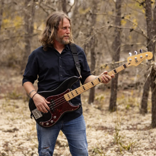 A man with long hair and a beard plays an electric bass guitar outdoors in a wooded area during autumn.