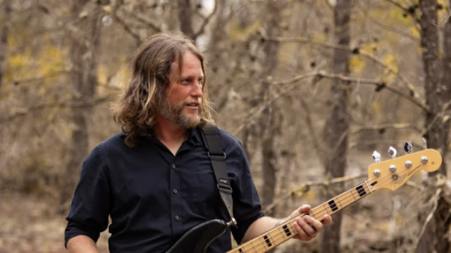 A man with long hair and a beard plays an electric bass guitar outdoors in a wooded area during autumn.