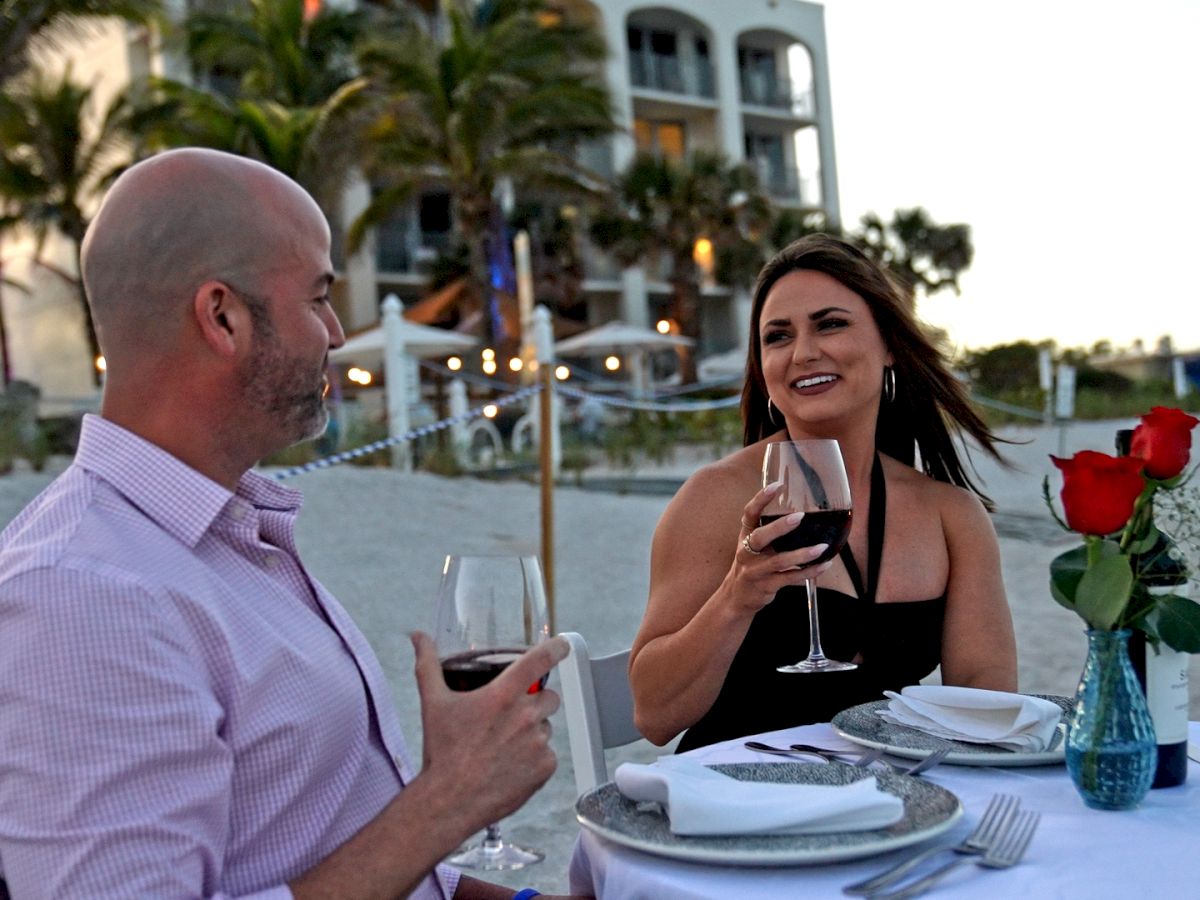 Two people are enjoying a seaside dinner, holding glasses of red wine, with a table set with plates and roses in a vase.