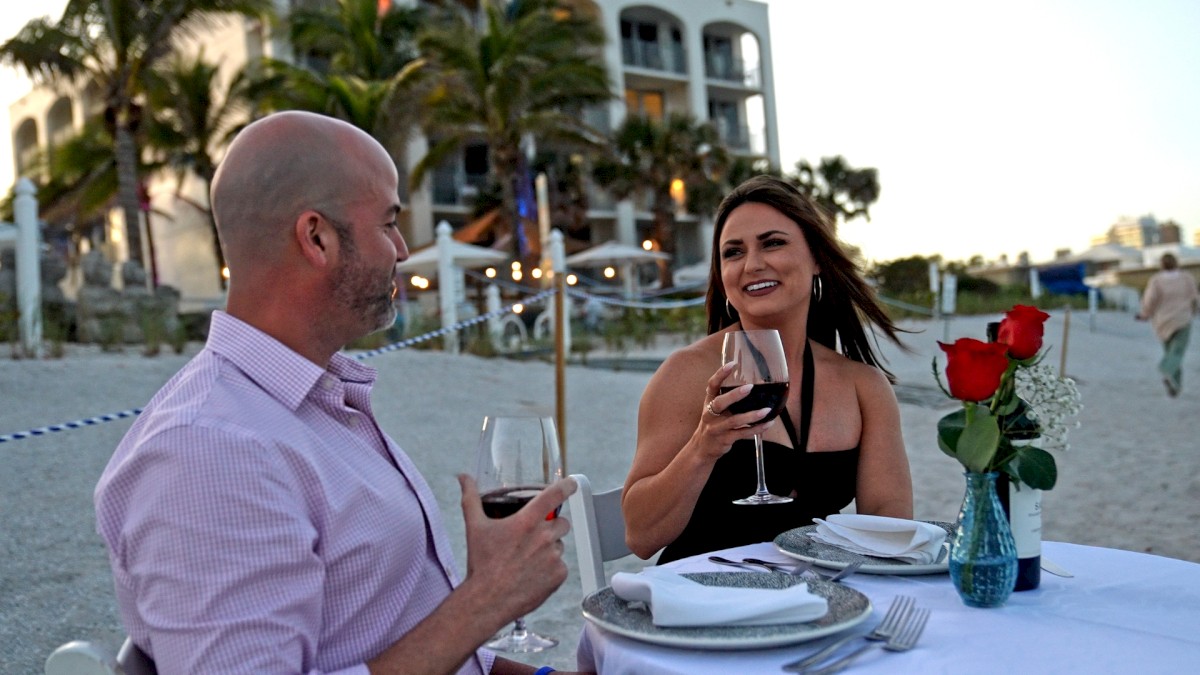 Two people are enjoying a seaside dinner, holding glasses of red wine, with a table set with plates and roses in a vase.