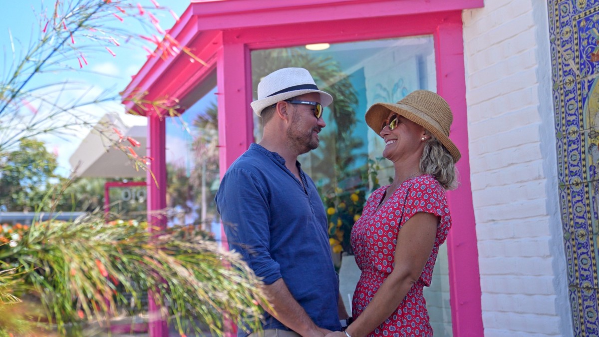 A couple stands in front of a building with pink trim, holding hands and smiling at each other, both wearing hats and sunglasses.