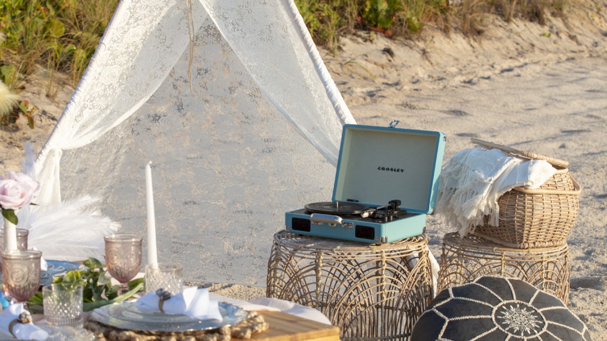 A beach setup features a teepee tent, a table with food, cushions, and a record player. The scene is set on the sand with greenery in the background.