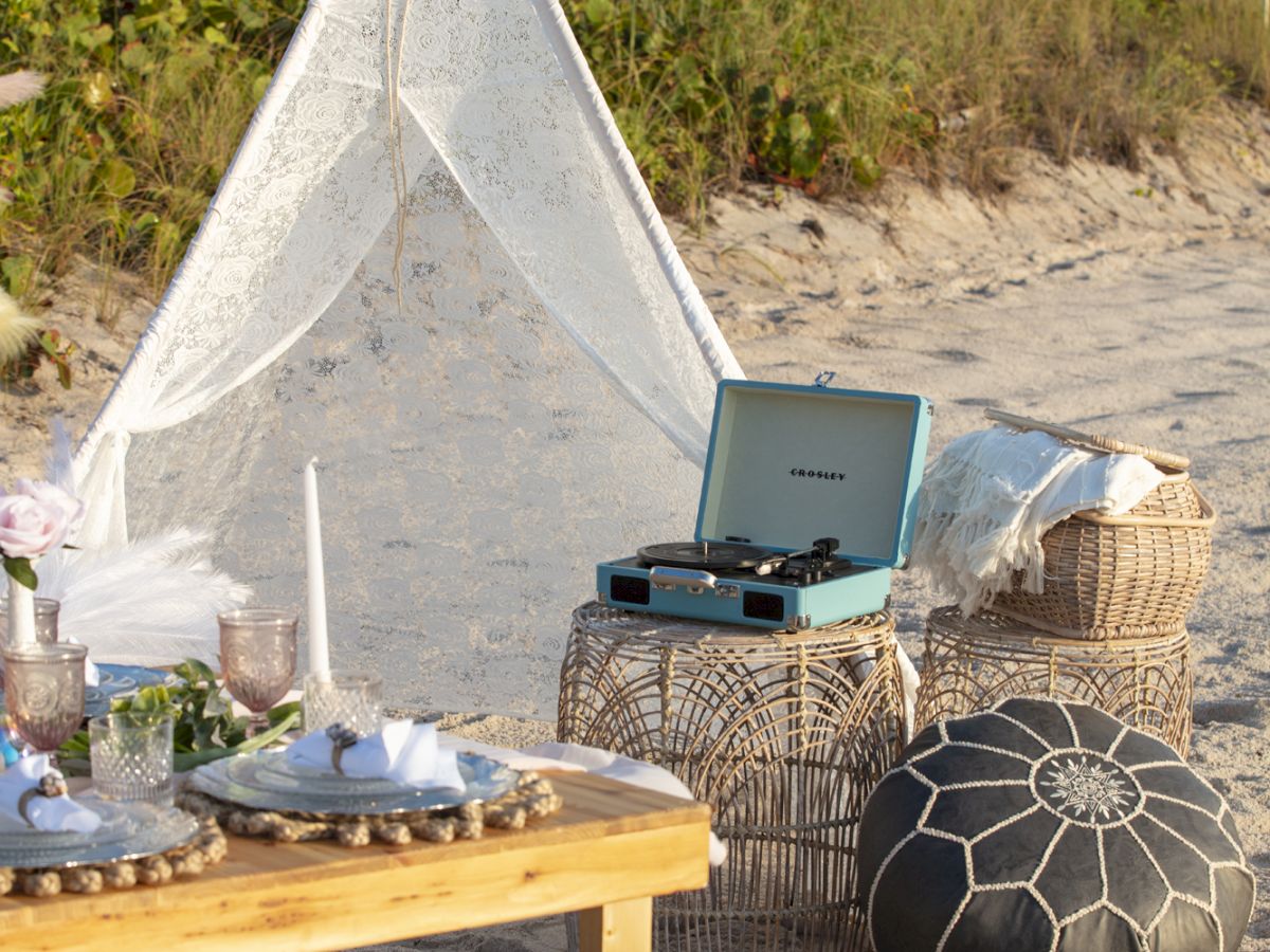 A beach setup features a teepee tent, a table with food, cushions, and a record player. The scene is set on the sand with greenery in the background.