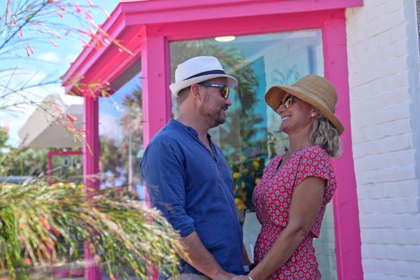 A couple in hats and sunglasses smile at each other, holding hands in front of a pink-trimmed building with a colorful, tiled wall.