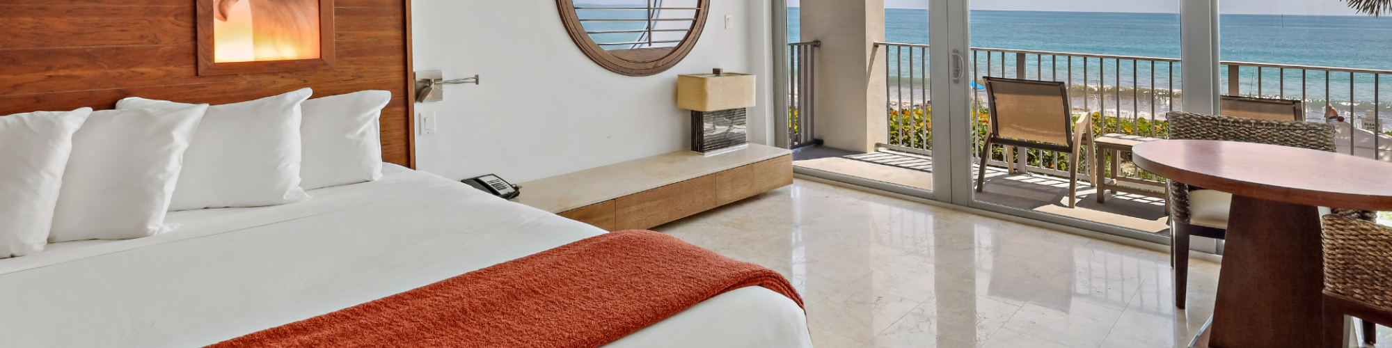 A modern hotel room features a large bed with an orange throw, a round mirror, a table with chairs, and a stunning ocean view through glass doors.