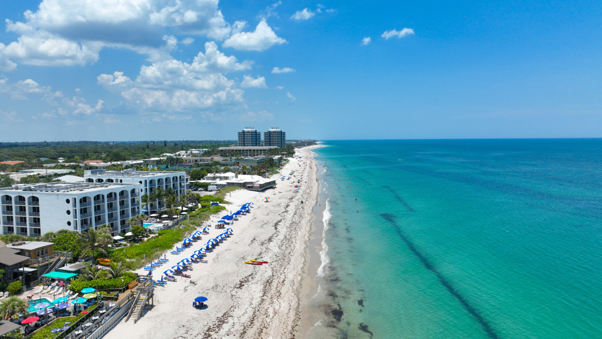 Aerial view of a beach with blue umbrellas, white buildings, and a clear ocean under a partly cloudy sky. People are visible on the beach.