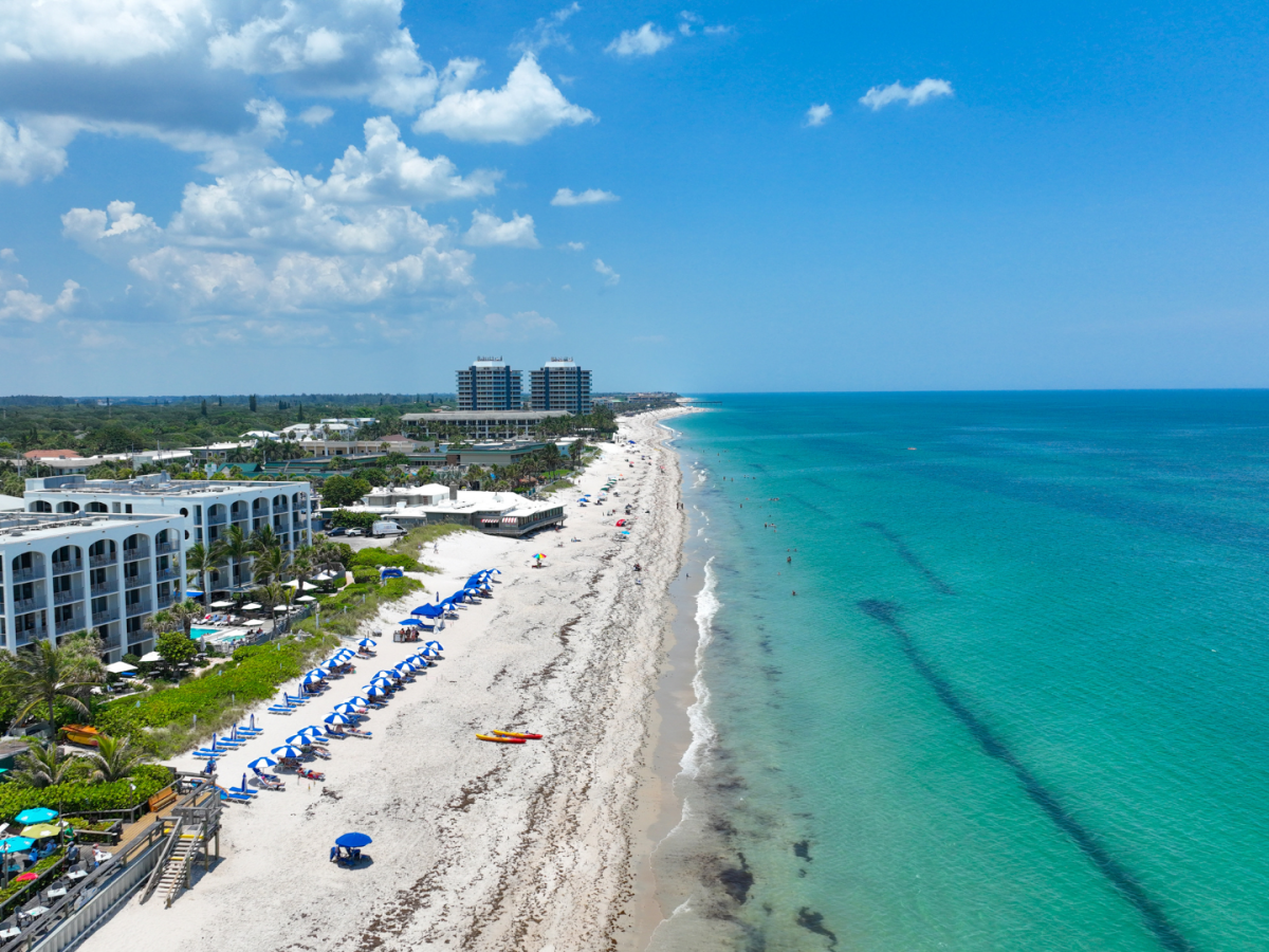 Aerial view of a beach with blue umbrellas, white buildings, and a clear ocean under a partly cloudy sky. People are visible on the beach.