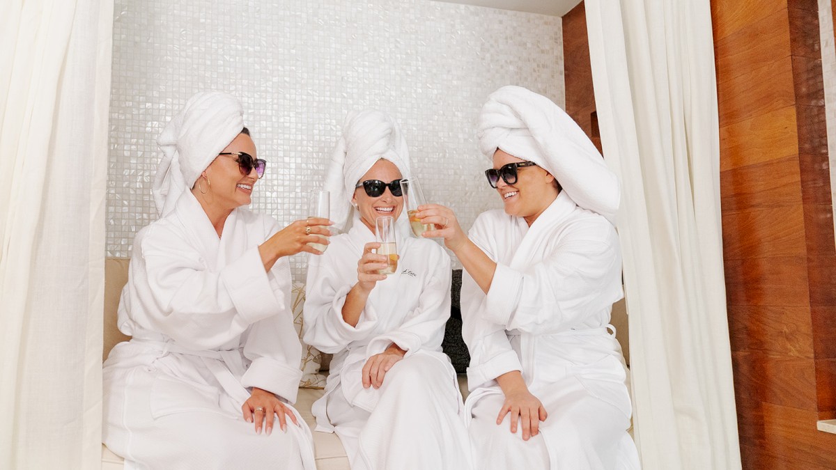 Three people in bathrobes with towels on their heads and wearing sunglasses, toasting with drinks in a relaxed setting.