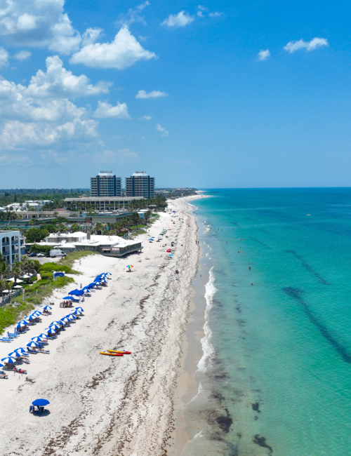 Aerial view of a beach with blue umbrellas, white buildings, and a clear ocean under a partly cloudy sky. People are visible on the beach.