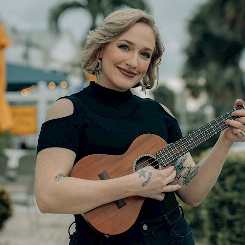 A woman with blonde hair and tattoos playing a ukulele outdoors, smiling at the camera with a blurred background of trees and sky.