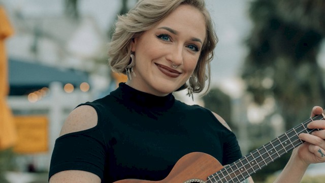 A woman with blonde hair and tattoos playing a ukulele outdoors, smiling at the camera with a blurred background of trees and sky.