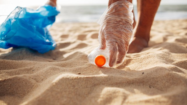 Someone is picking up litter, specifically a plastic bottle, from the beach to help keep it clean and protect the environment.