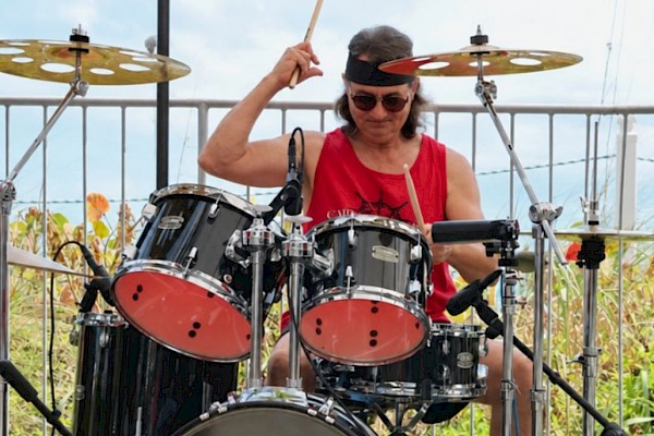 A woman playing a drum set outdoors, wearing sunglasses and a red tank top, with 
