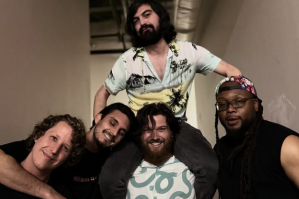 A group of five men, one sitting on others, smiling and posing together in a casual setting.