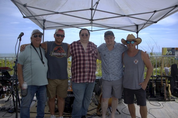 Five band members posing together on an outdoor stage with musical instruments and equipment in the background.