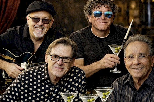 Four musicians posing with guitars and martini glasses in a relaxed group photo.