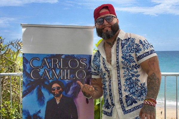 ai description Musician wearing a blue-and-white floral outfit posing beside a Carlos Camilo promotional banner, with the ocean in the background.