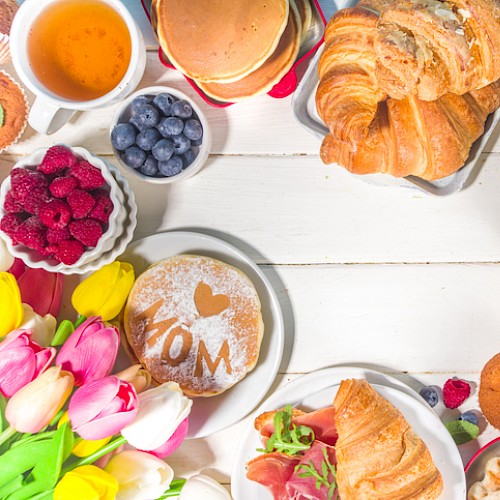 A breakfast spread with croissants, eggs, berries, muffins, honey, flowers, and beverages on a white table.