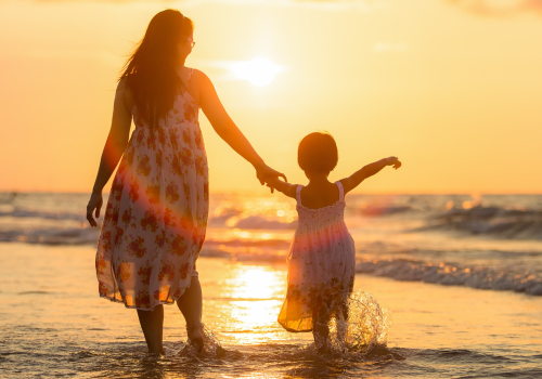 A woman and a child hold hands while walking along the beach at sunset, with waves gently touching their feet and the sky glowing warm hues of orange.
