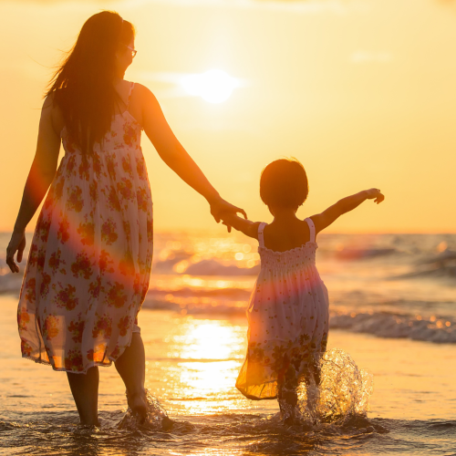 A woman and a child hold hands while walking along the beach at sunset, with waves gently touching their feet and the sky glowing warm hues of orange.