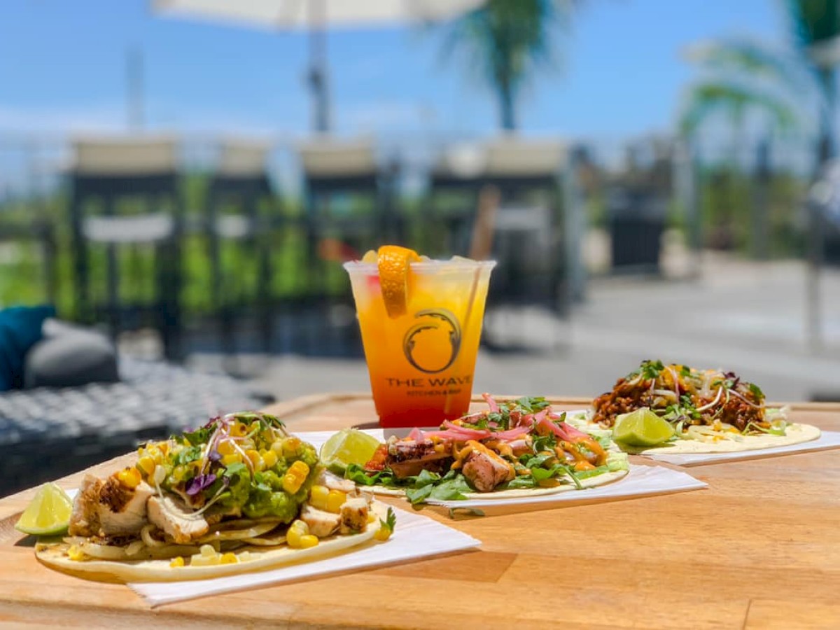 An outdoor table with three loaded tacos, lime wedges, and a colorful drink in a cup labeled 