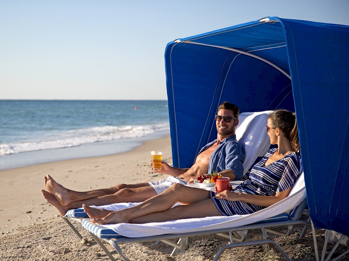 Two people are relaxing on lounge chairs under a blue canopy at the beach, enjoying drinks and smiling at each other near the shoreline.