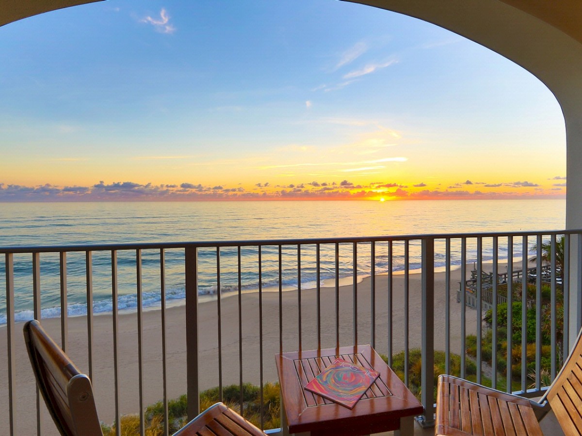 A serene beach view from a balcony at sunset, featuring two wooden chairs and a small table, with the sun setting over the horizon in the background.