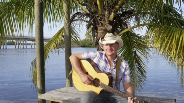 A man in a plaid shirt and cowboy hat plays guitar near a lake under a palm tree.