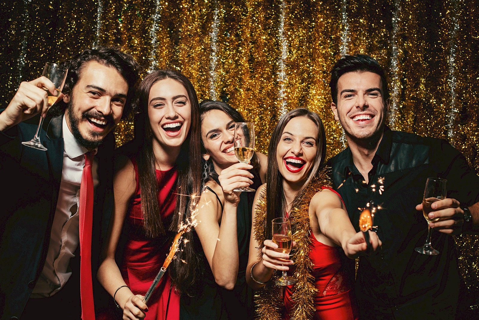 A group of friends celebrate at a party, wearing festive attire and holding drinks, with a sparkler and a gold backdrop.