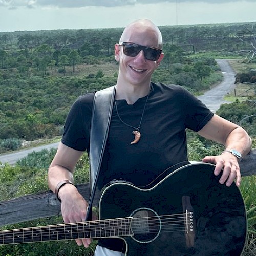 A person with sunglasses stands by a fence, holding a black guitar in a scenic outdoor area.