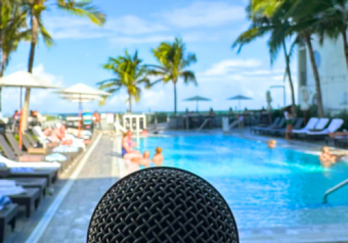 A microphone is in the foreground, with people relaxing by a pool surrounded by palm trees under a clear blue sky.