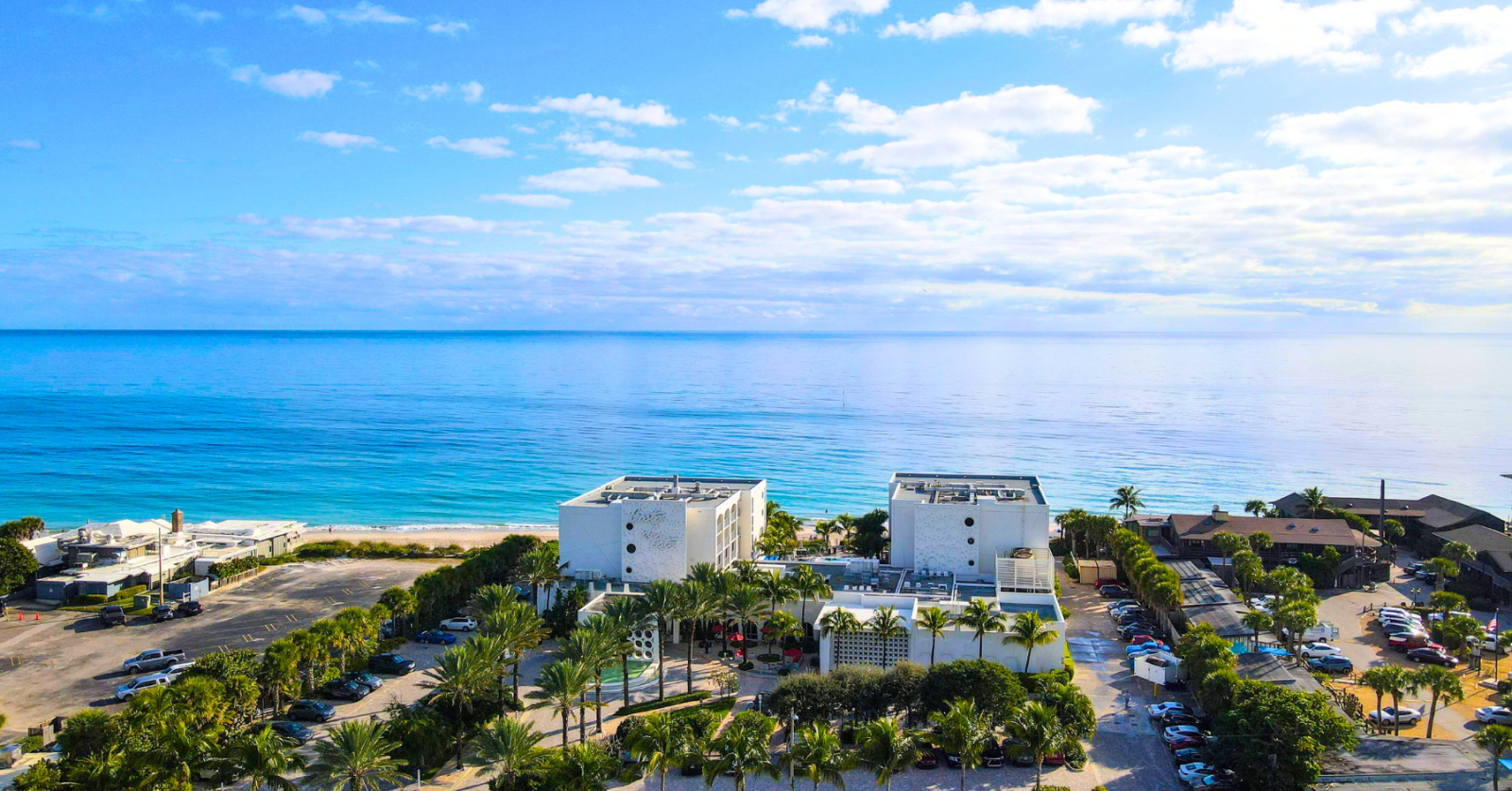 Aerial view of a beachfront area with buildings, palm trees, parked cars, and a calm ocean under a blue sky.
