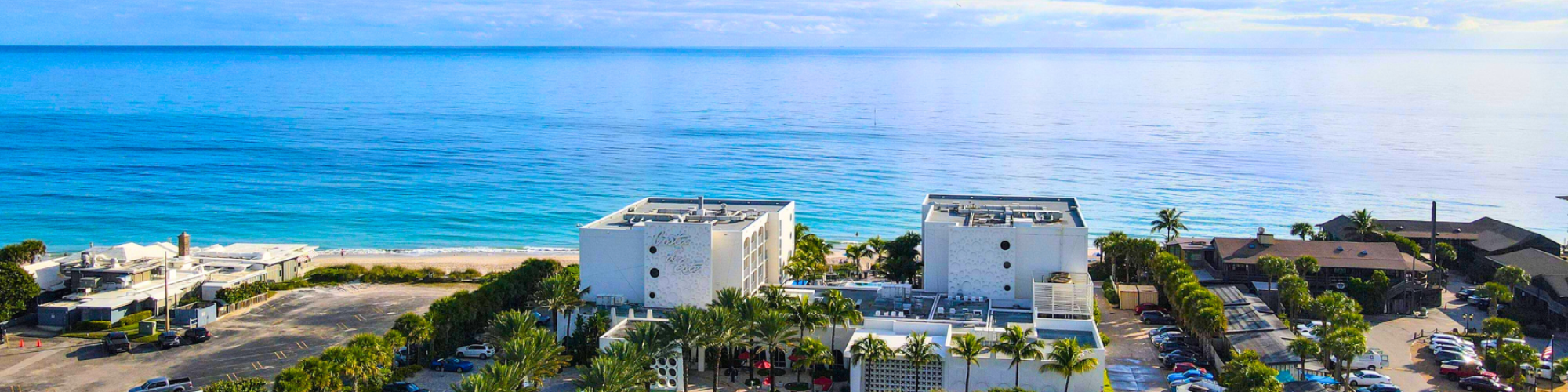 Aerial view of a beachfront area with buildings, palm trees, parked cars, and a calm ocean under a blue sky.