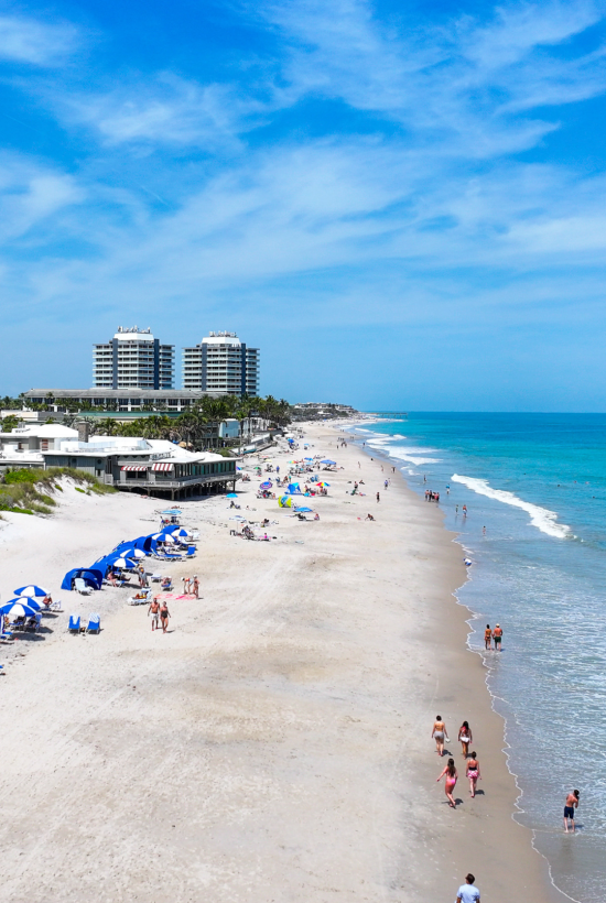 A sunny beach scene with several people, umbrellas, and buildings in the background.