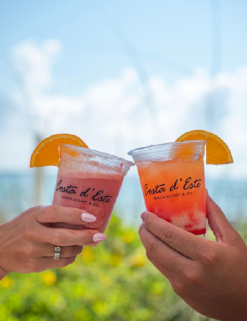 Two hands hold colorful drinks with orange slices, celebrating outdoors under a sunny, blue sky with clouds, at a beach resort.