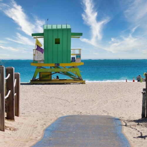 A sandy beach with a colorful lifeguard tower, wooden posts, and roped fences leading to the ocean, under a bright blue sky with wispy clouds.