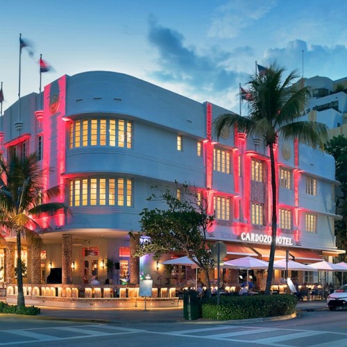 This image shows an art deco style hotel with pink neon lights, palm trees, and outdoor seating on a street corner during the evening.