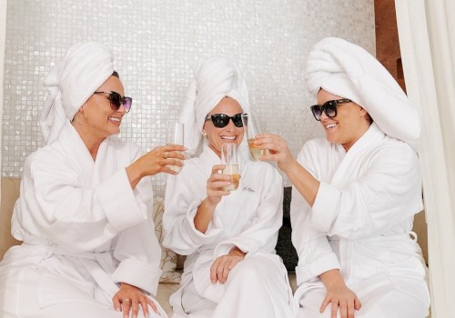 Three people in white bathrobes and towels on their heads, wearing sunglasses, are sitting and toasting with champagne glasses in a relaxed setting.