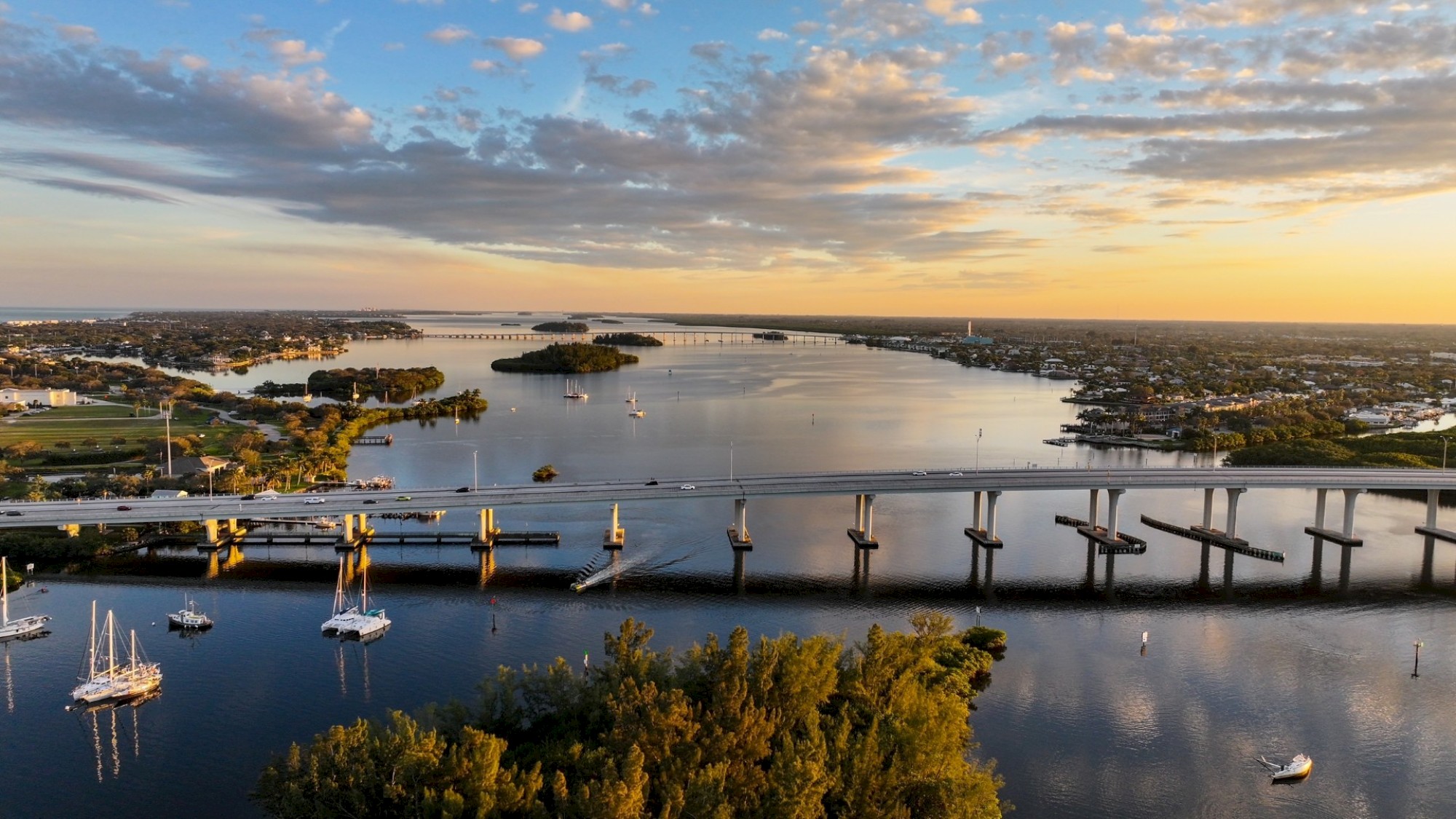 An aerial view of a scenic waterway at sunset with boats and bridges spanning the water, surrounded by greenery and small islands.