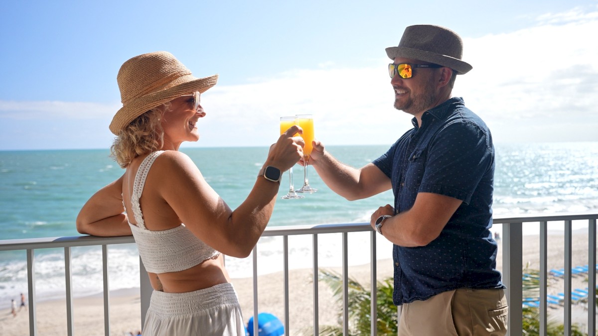 Two people in hats clink glasses on a balcony with an ocean view, smiling and dressed in casual summer attire, enjoying a sunny day.