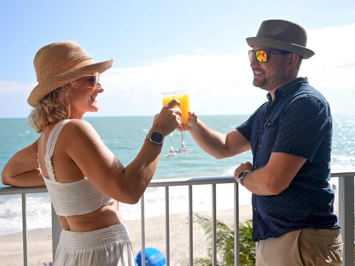 Two people in hats clink glasses on a balcony with an ocean view, smiling and dressed in casual summer attire, enjoying a sunny day.