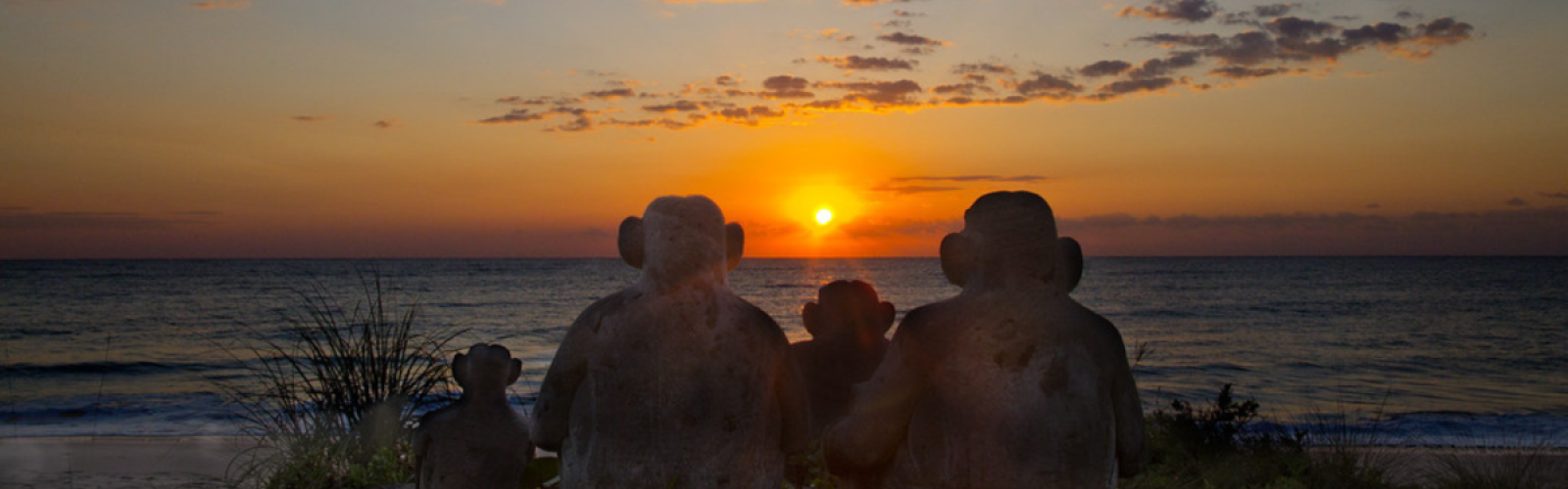 Stone sculptures of monkeys sit facing a sunset over the ocean at the beach.