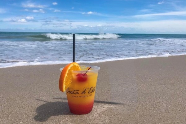 A colorful cocktail with an orange slice on a sandy beach, with the ocean and blue sky in the background.