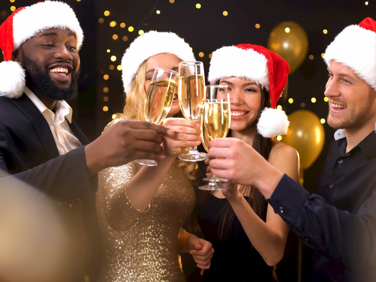 A group of people wearing Santa hats clink champagne glasses in a festive celebration with gold balloons in the background.