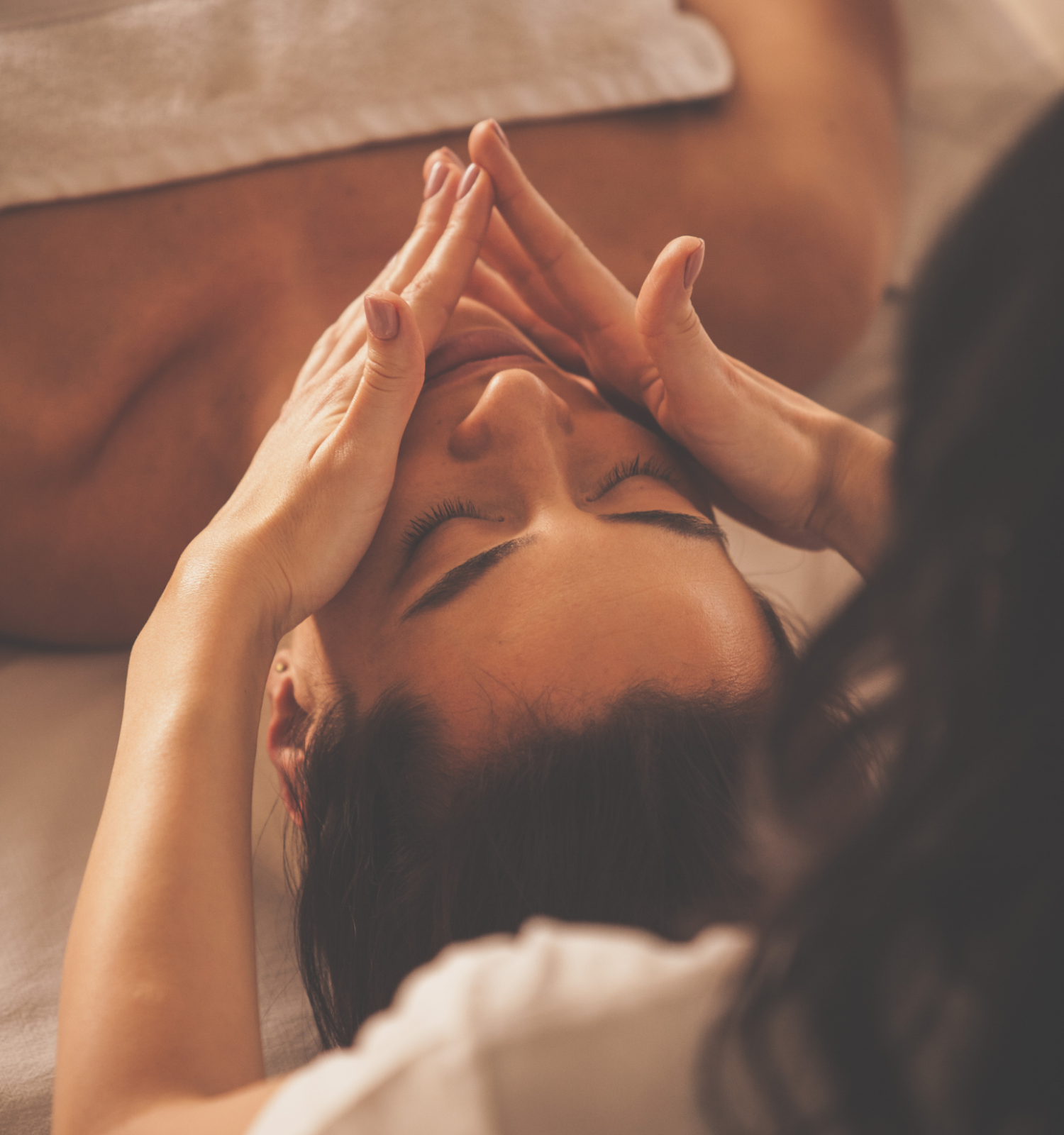 A person is receiving a facial treatment while lying down, covered partially by a towel, with another person's hands gently massaging their face.