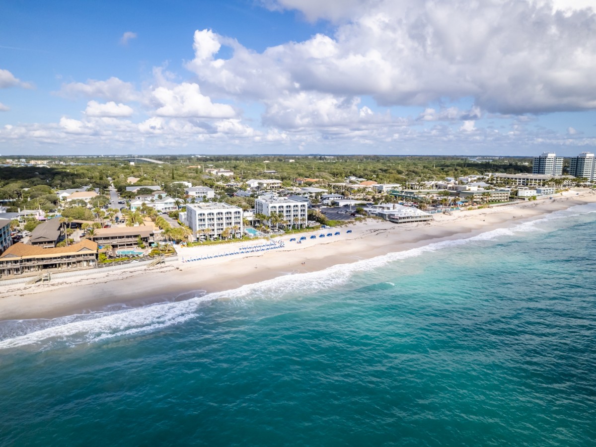 Aerial view of a coastal town with sandy beaches, clear blue water, buildings, and green landscape under a partly cloudy sky.