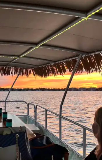 Two people on a covered boat enjoying a sunset on the water, with drinks and a phone on the table.