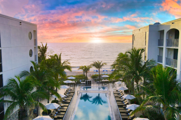 A stunning ocean view between two buildings, with a pool surrounded by palm trees and lounge chairs, set against a colorful sunset.