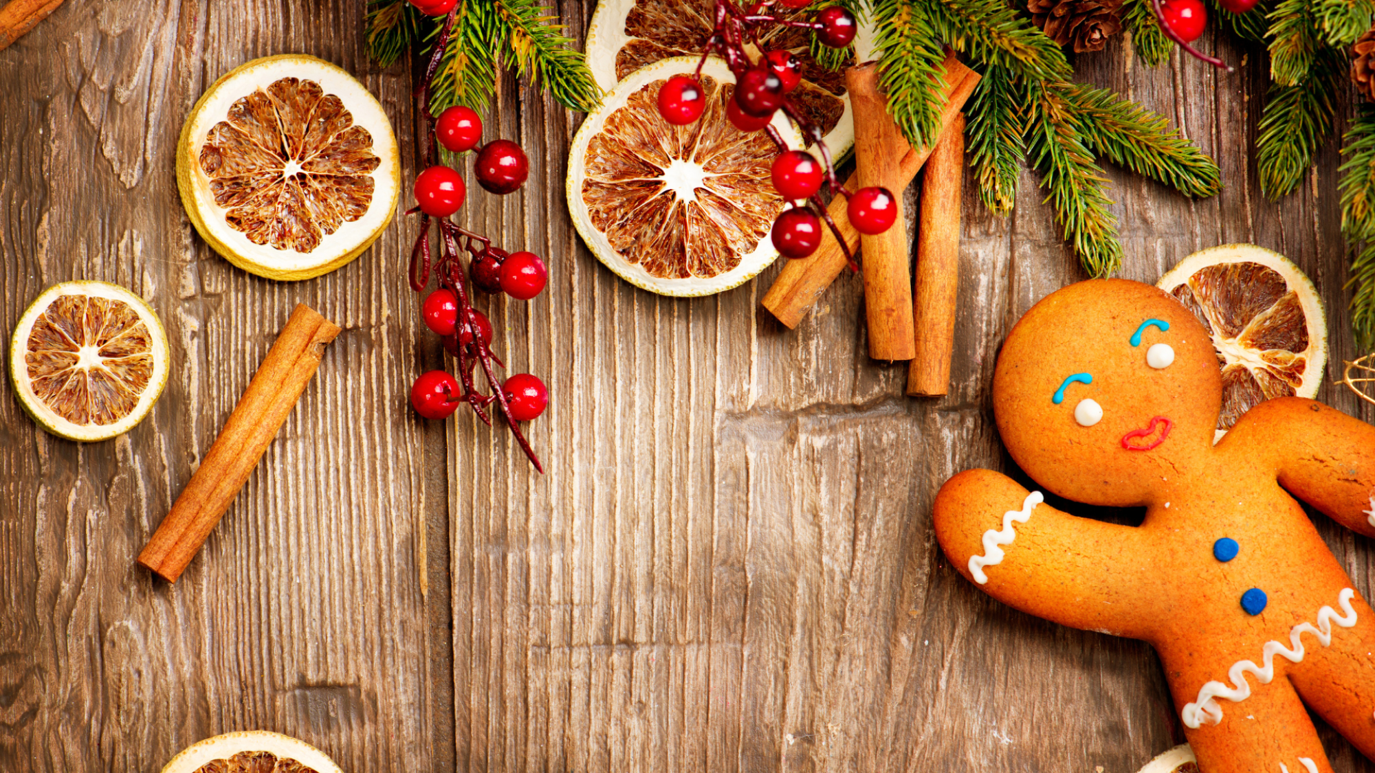 A wooden surface is decorated with dried orange slices, cinnamon sticks, red berries, green pine branches, and a smiling gingerbread cookie.